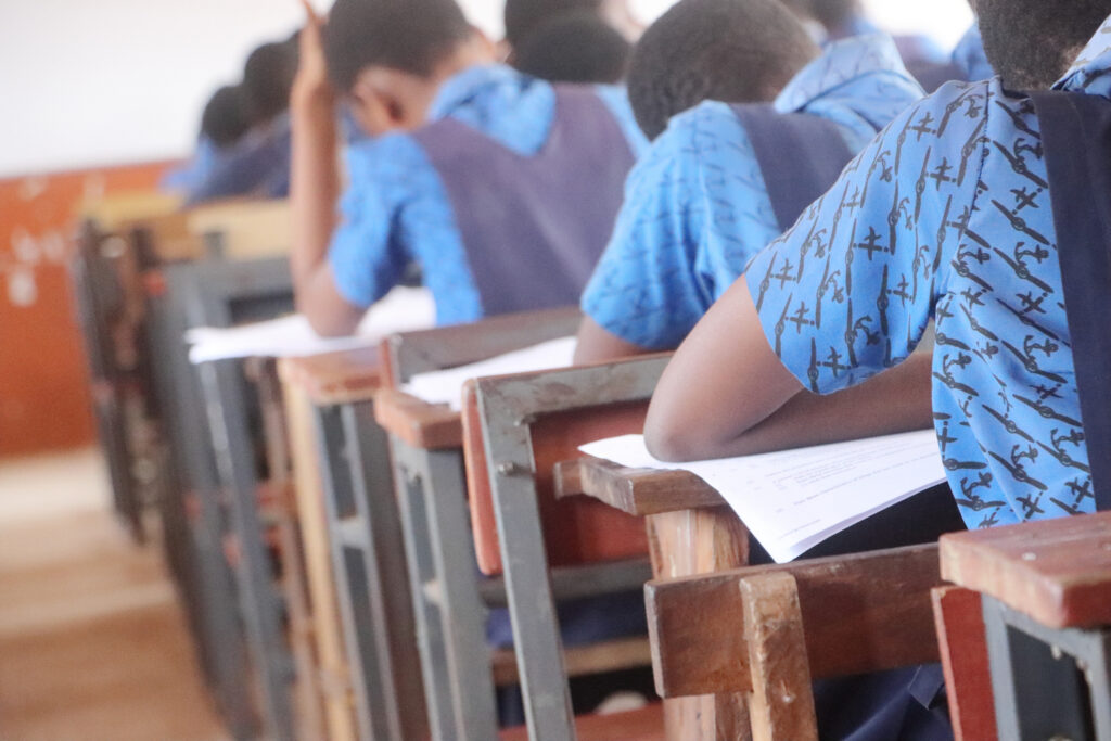 An intimate perspective on the crucial Basic Education Certificate Examination (BECE) in Ghana, featuring a close-up of a student's hand poised with a pen, actively engaged in writing. The subtle blur of classmates in the background underscores the shared, serious atmosphere of the examination. The distinct Ghanaian public or private school uniform worn by the student grounds the image firmly in its authentic West African setting. This visual encapsulates the dedication of young Ghanaians striving for educational advancement and the future generation's pursuit of knowledge in Africa.