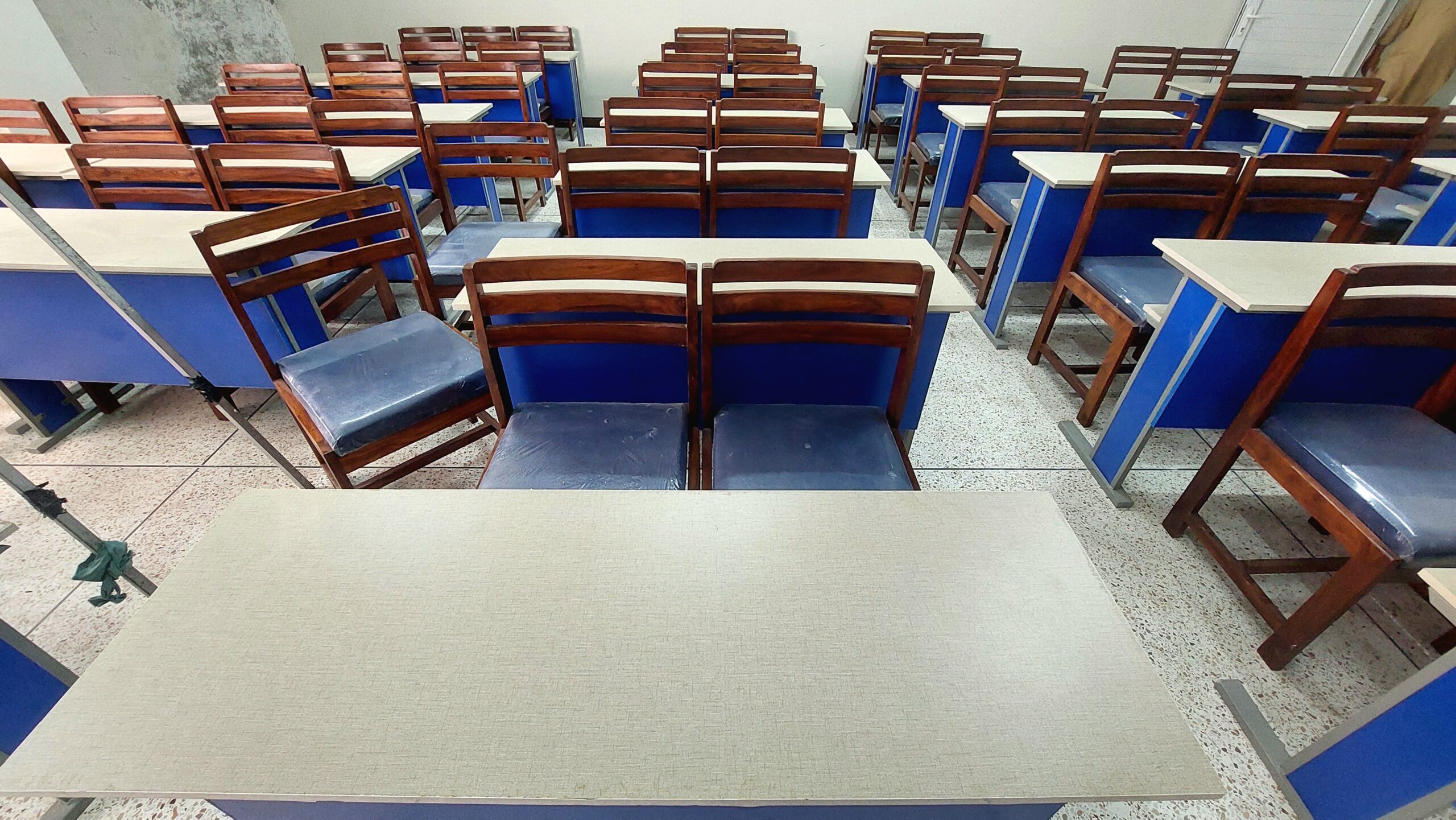 Classroom interior featuring neatly arranged wooden desks and chairs with blue accents and cushioned seats. The setting is well-lit, highlighting the uniform arrangement and orderly appearance. The flooring is a speckled tile, adding to the bright and clean atmosphere. Desks are aligned in multiple rows, suggesting a traditional educational environment. Overall, the space appears organized and conducive to learning activities.